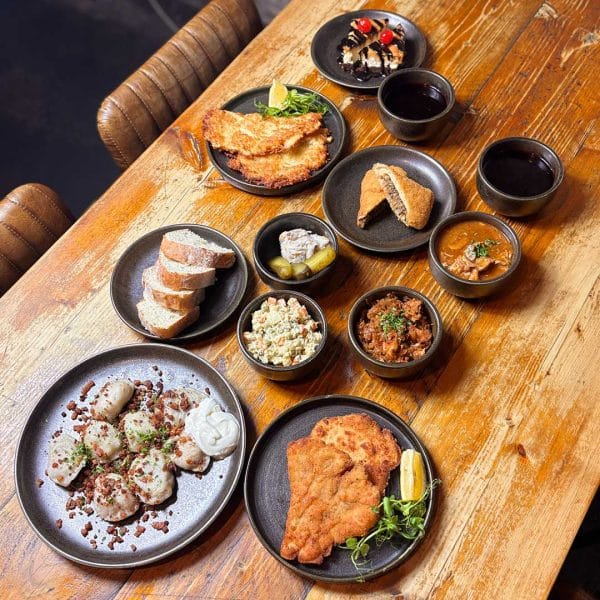 A long wooden table set with multiple plates of comfort foods: fried chicken, bread slices, potato salad, pickles, bowls of curry and stews, and a dessert plate in the back corner.