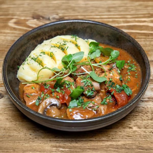 Beef and mushroom stew in a brown bowl with red peppers and tomato sauce, topped with sliced potatoes and fresh herbs.
