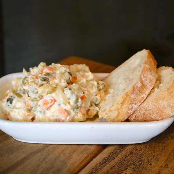 Creamy potato salad with diced carrots, pickles, and celery on a white plate, with crusty bread beside it.
