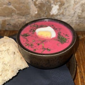Bright pink beet soup (borscht) in a rustic bowl, topped with a halved hard-boiled egg and chopped dill, with crusty bread on the side