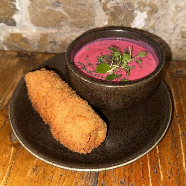 Bowl of bright pink beet soup with fresh herbs, served with a crispy breaded croquette on a dark plate.