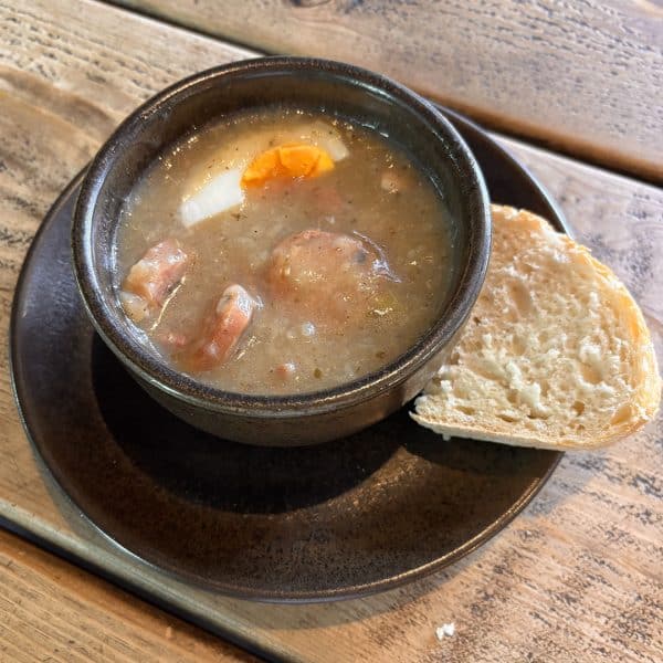 Creamy soup with chunks of meat, carrot, and onion in a dark bowl, served with crusty bread on a wooden table.