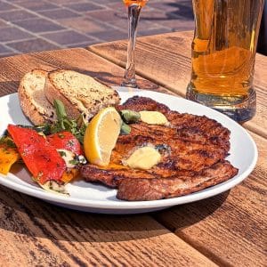 Plate with grilled steak, lemon wedge, a dollop of sauce, roasted vegetables, and crusty bread on a wooden table, with beer and a glass in the background.