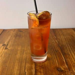 Tall highball glass of iced tea with lime and orange slices, and a black straw, on a wooden table.