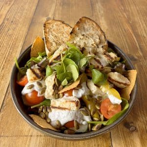 Bowl of salad with arugula, cherry tomatoes, dollops of creamy cheese, toasted bread pieces, and pumpkin seeds on a wooden table.