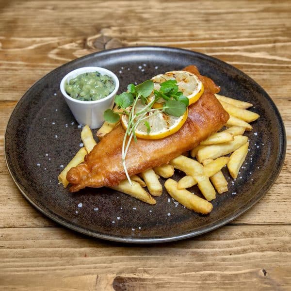 Golden fried fish fillet on a bed of French fries, topped with lemon slices and microgreens, with a side of green dipping sauce.