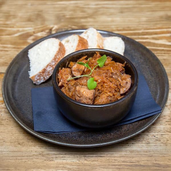 Braised shredded meat with caramelized onions in a black bowl, garnished with greens, served with slices of crusty bread on a dark plate.