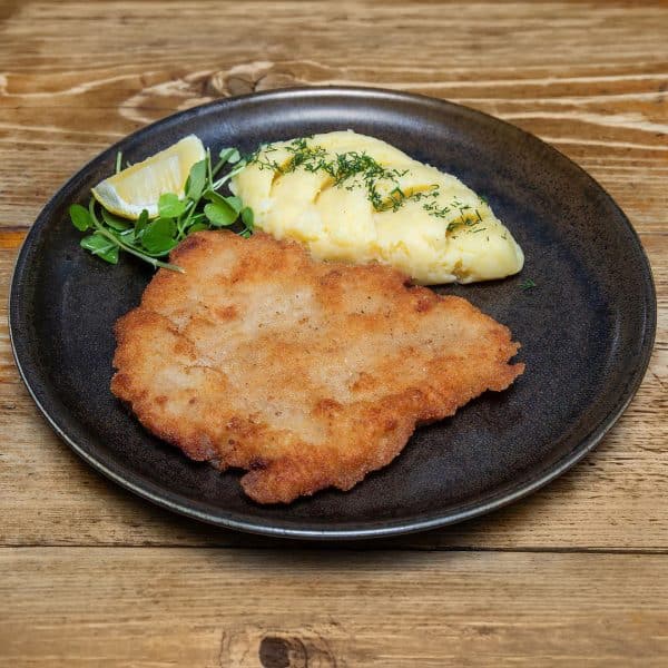 Crispy breaded schnitzel on a plate with mashed potatoes, lemon wedge, and greens on a dark plate.
