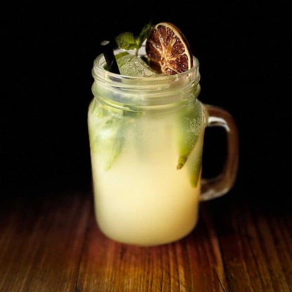 Mason jar of pale mojito with ice, lime wedges, and mint, on a dark wooden surface.