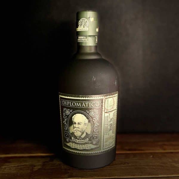 Diplomático Reserva Exclusiva rum bottle with ornate label and portrait on a dark wooden table outside a black background.