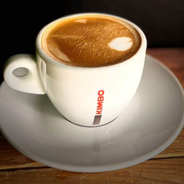 White ceramic cup of coffee with crema on top, placed on a light gray saucer on a wooden table; red KIMBO branding on the cup.
